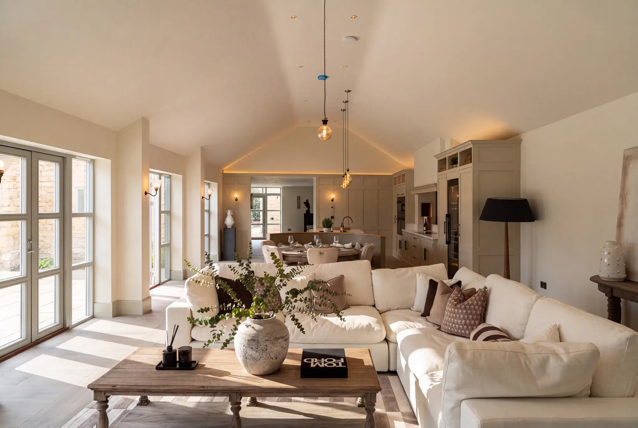 An open-plan living room and kitchen under a vaulted ceiling, with pendant lights, wall lights between the windows and a line of concealed lighting tracing the gable
