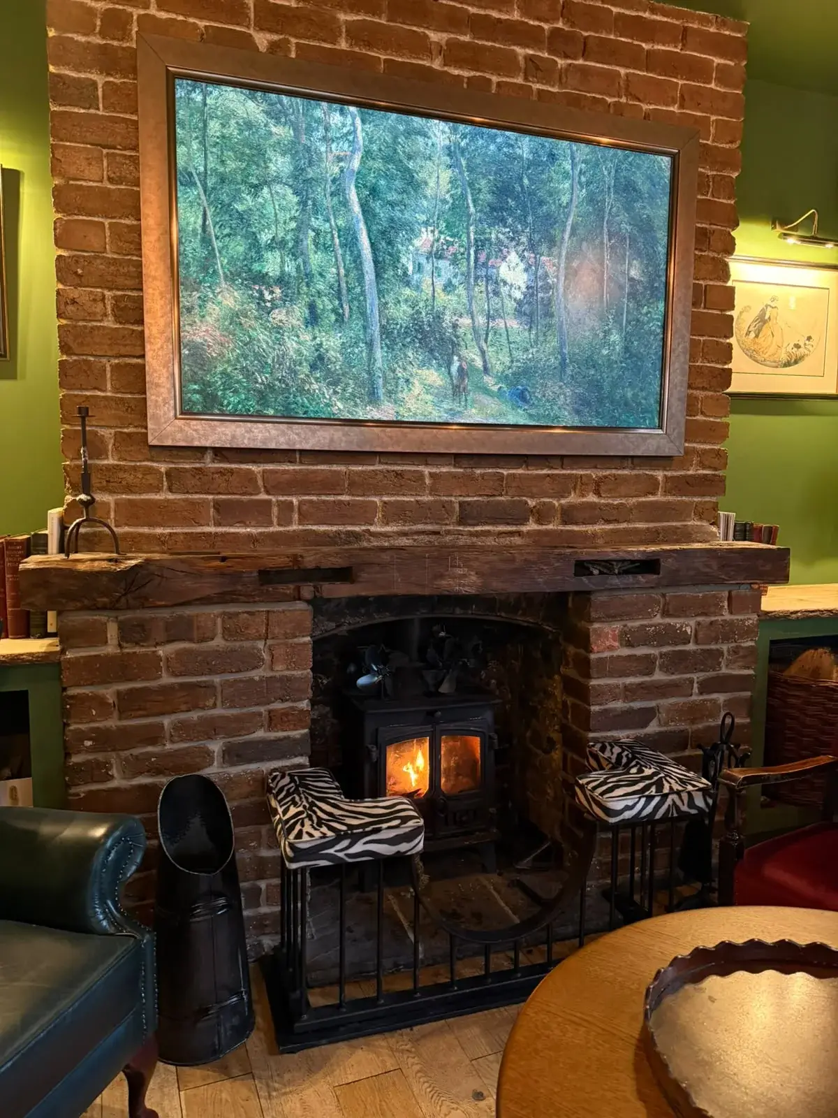 A wall-mounted screen showing a painting of a woodland path, hung on an exposed brick chimney breast above a lit wood burner