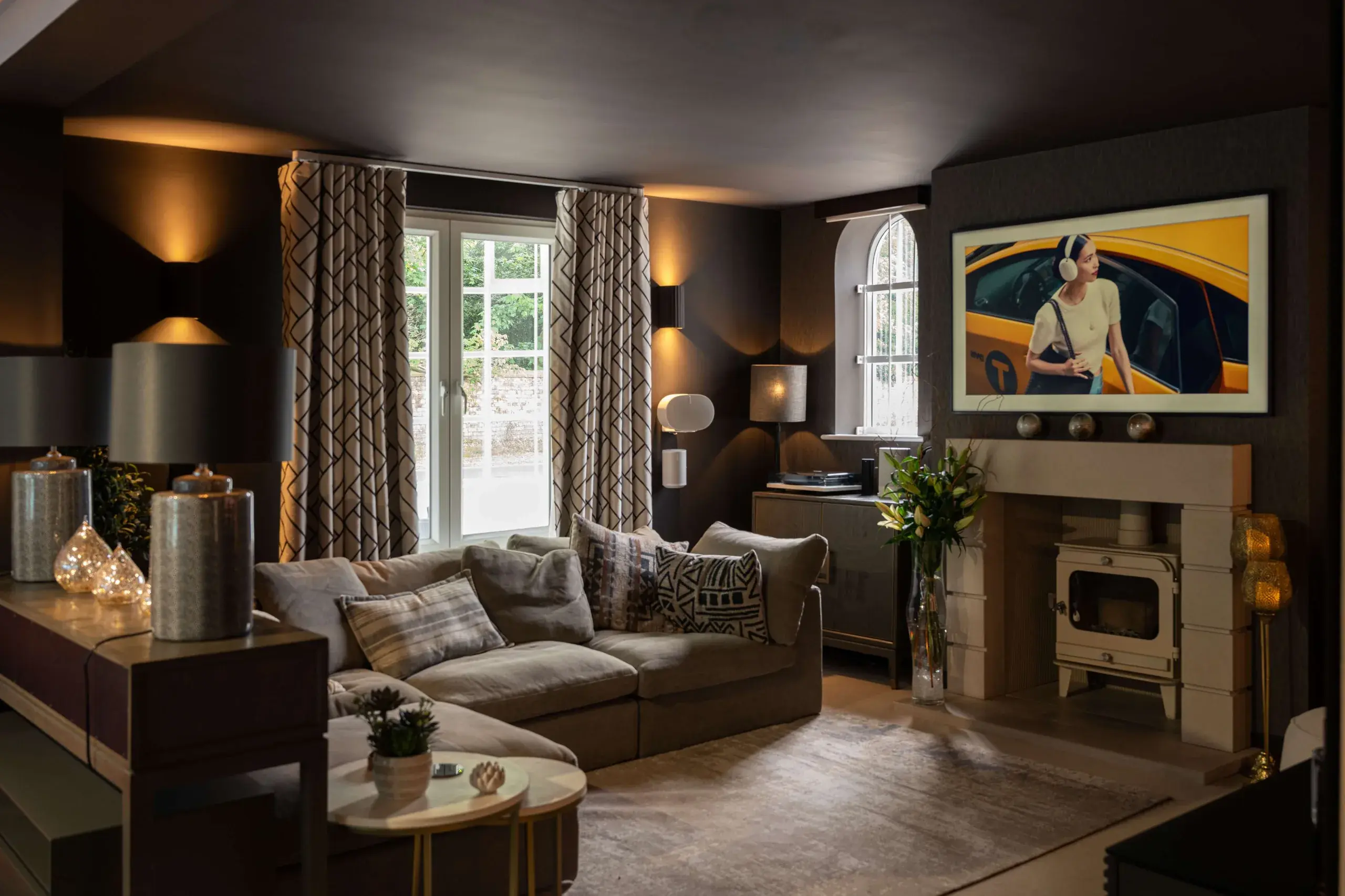The showroom living room, with a wood burner, a wall-mounted television displaying a picture, and layered lighting